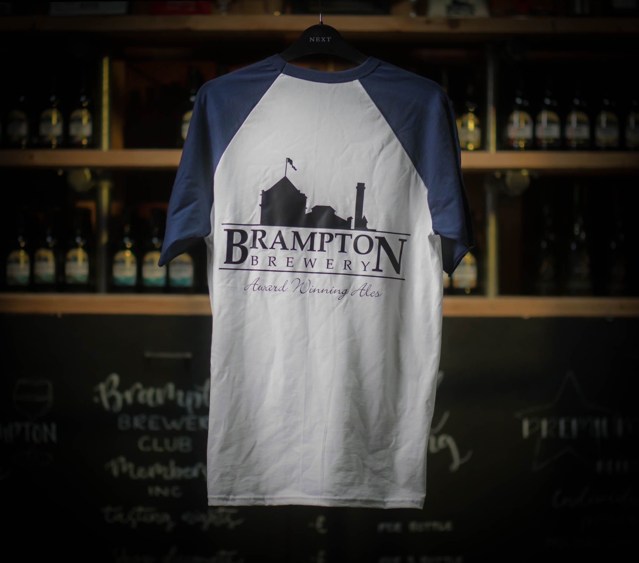 White and navy baseball Brampton Brewery T‑shirt hanging in a rustic brewery setting, featuring the brewery’s logo and “Award Winning Ales” text.