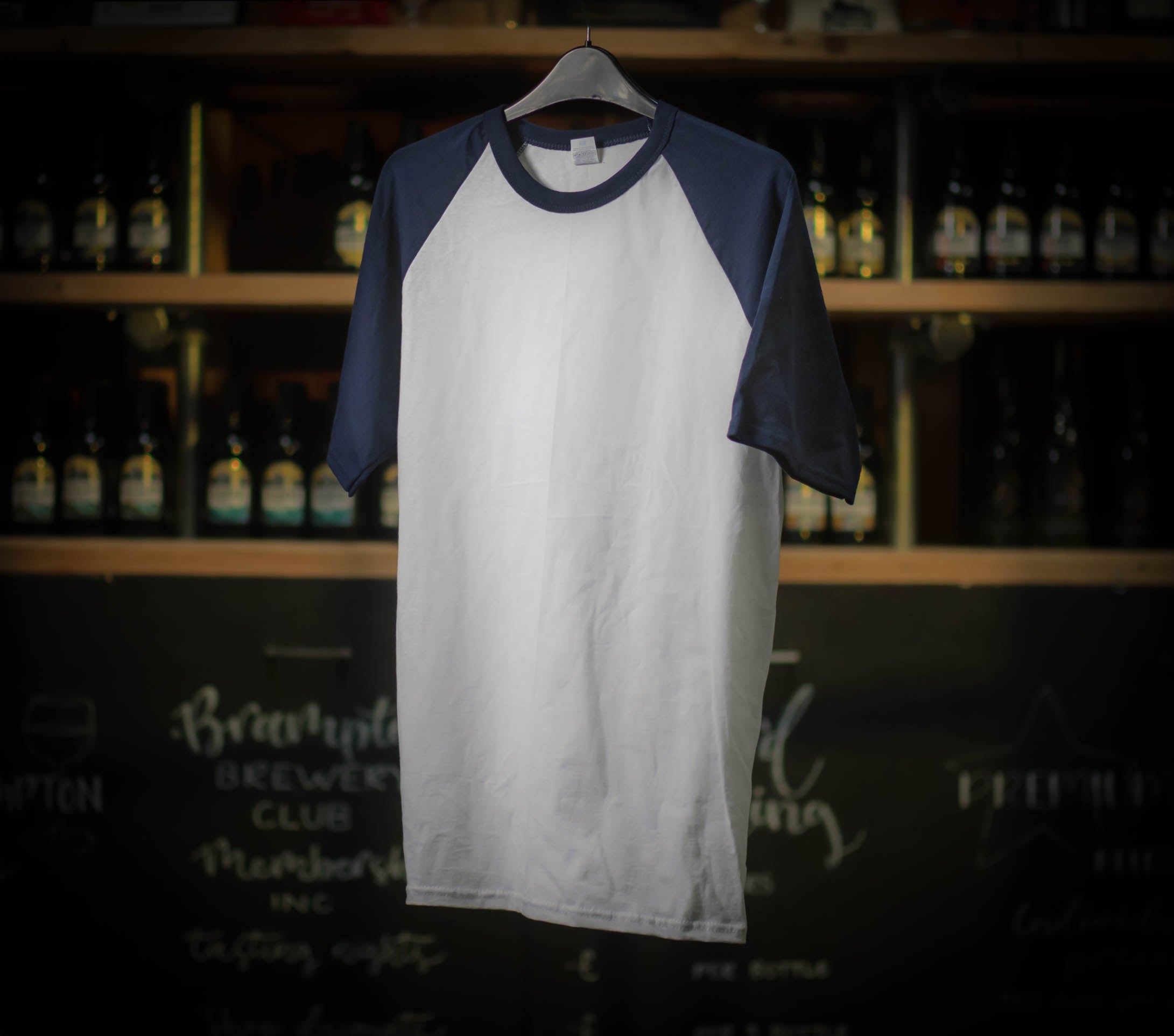 White and navy baseball Brampton Brewery T‑shirt hanging in a rustic brewery setting.