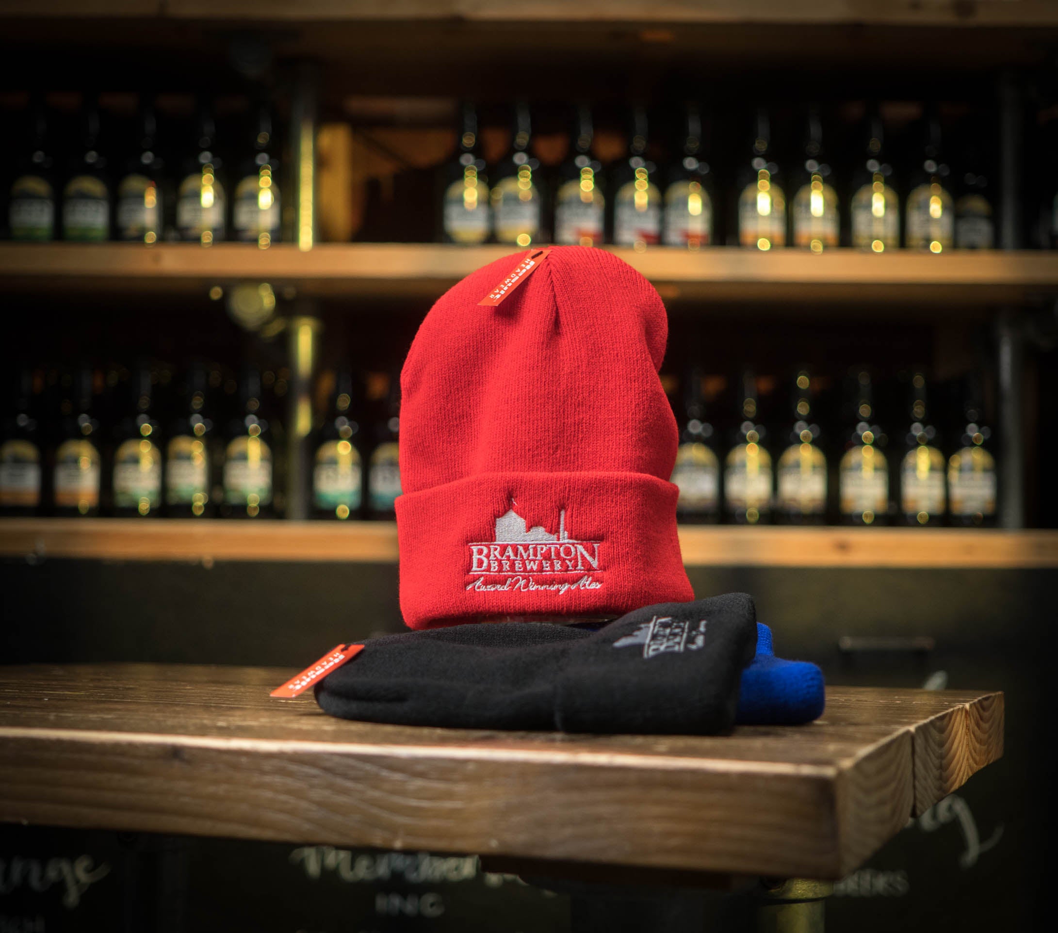 Red beanie with a Brampton Brewery logo on a wooden surface with bottles in the background