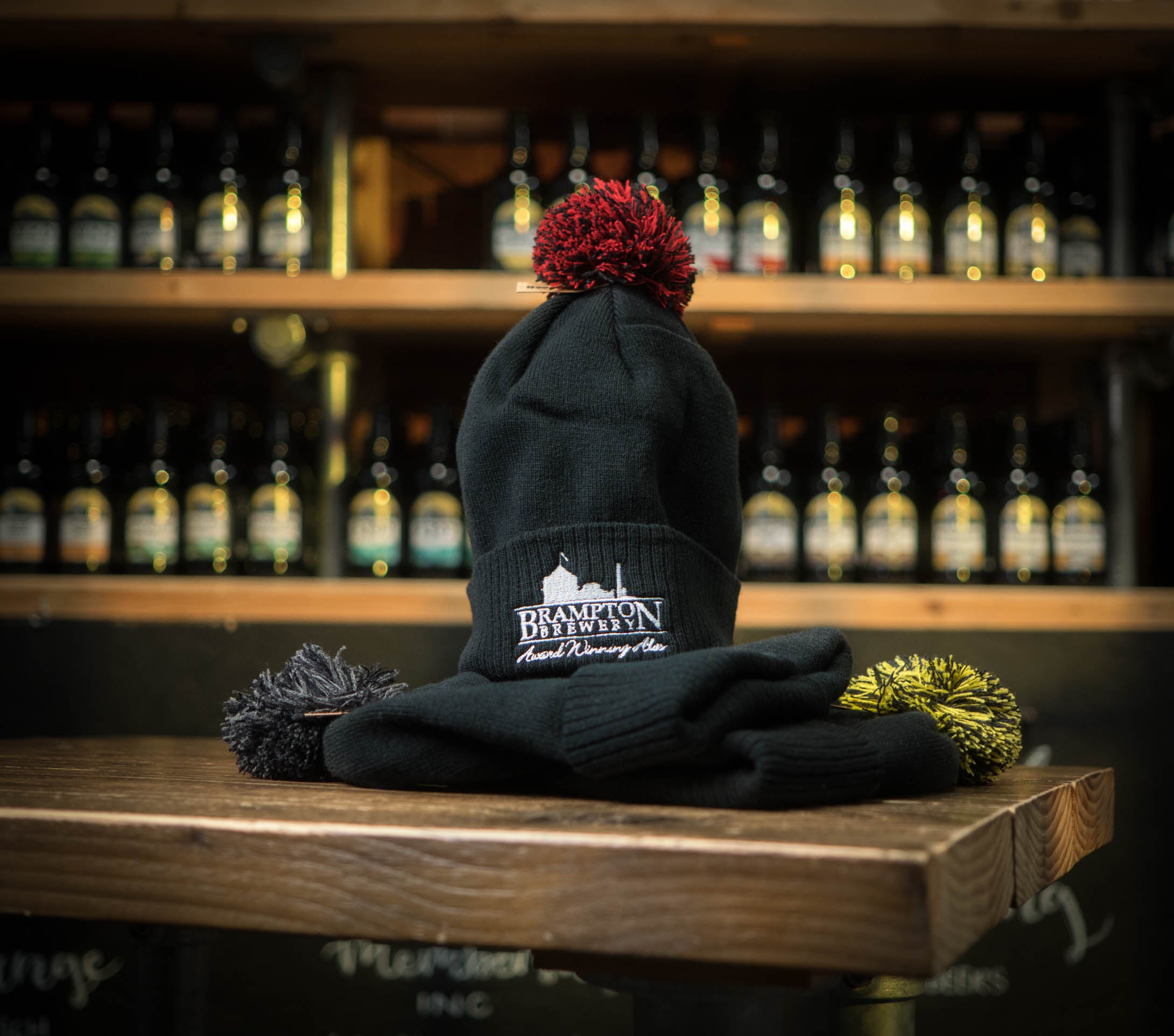 Black beanie with red pom-pom and a Brampton Brewery logo on a wooden surface with bottles in the background