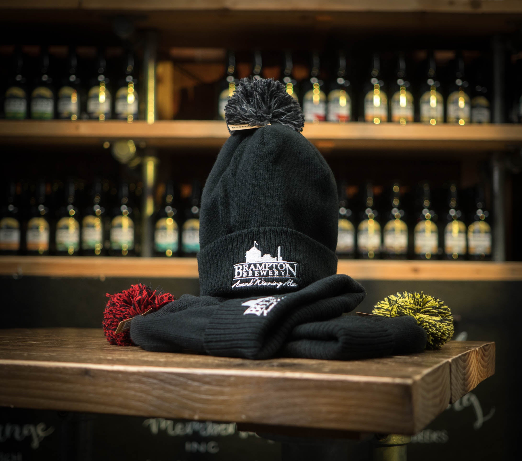Black beanie with black pom-pom and Brampton Brewery logo on a wooden surface with bottles in the background