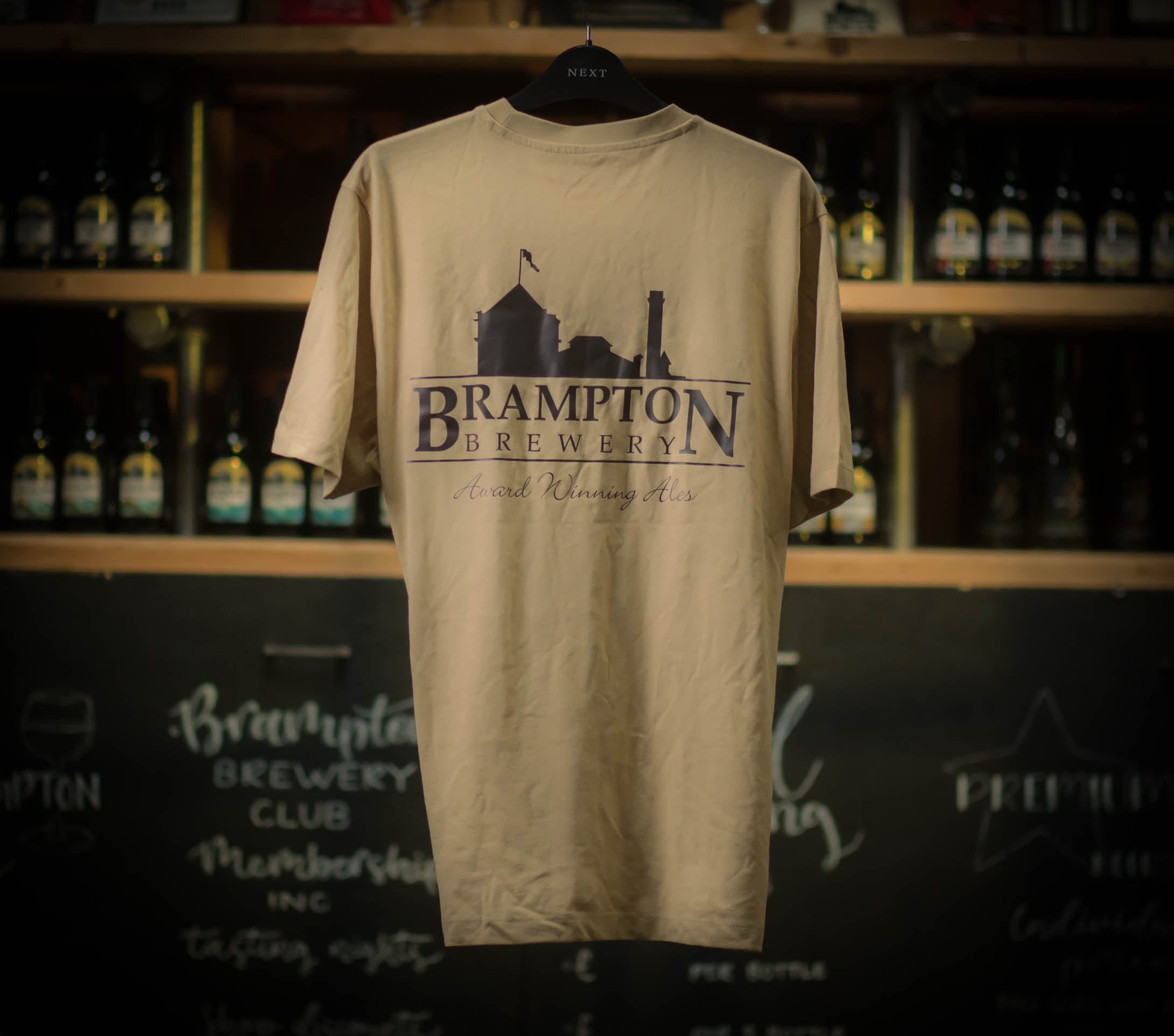 Beige Brampton Brewery T‑shirt hanging in a rustic brewery setting, featuring the brewery’s logo and “Award Winning Ales” text.