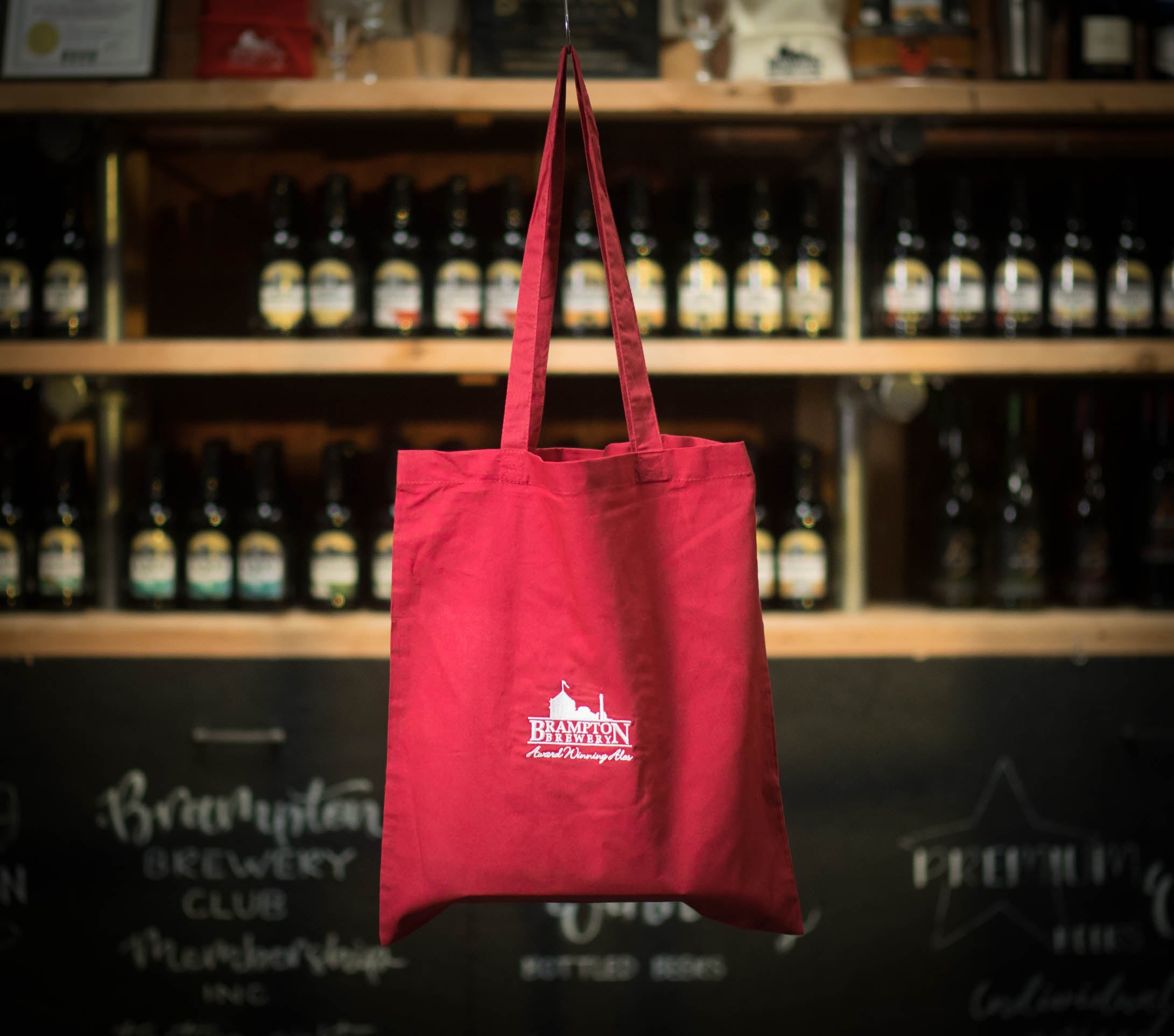 Red tote bag with a Brampton Brewery logo hanging in front of a shelf of bottles.