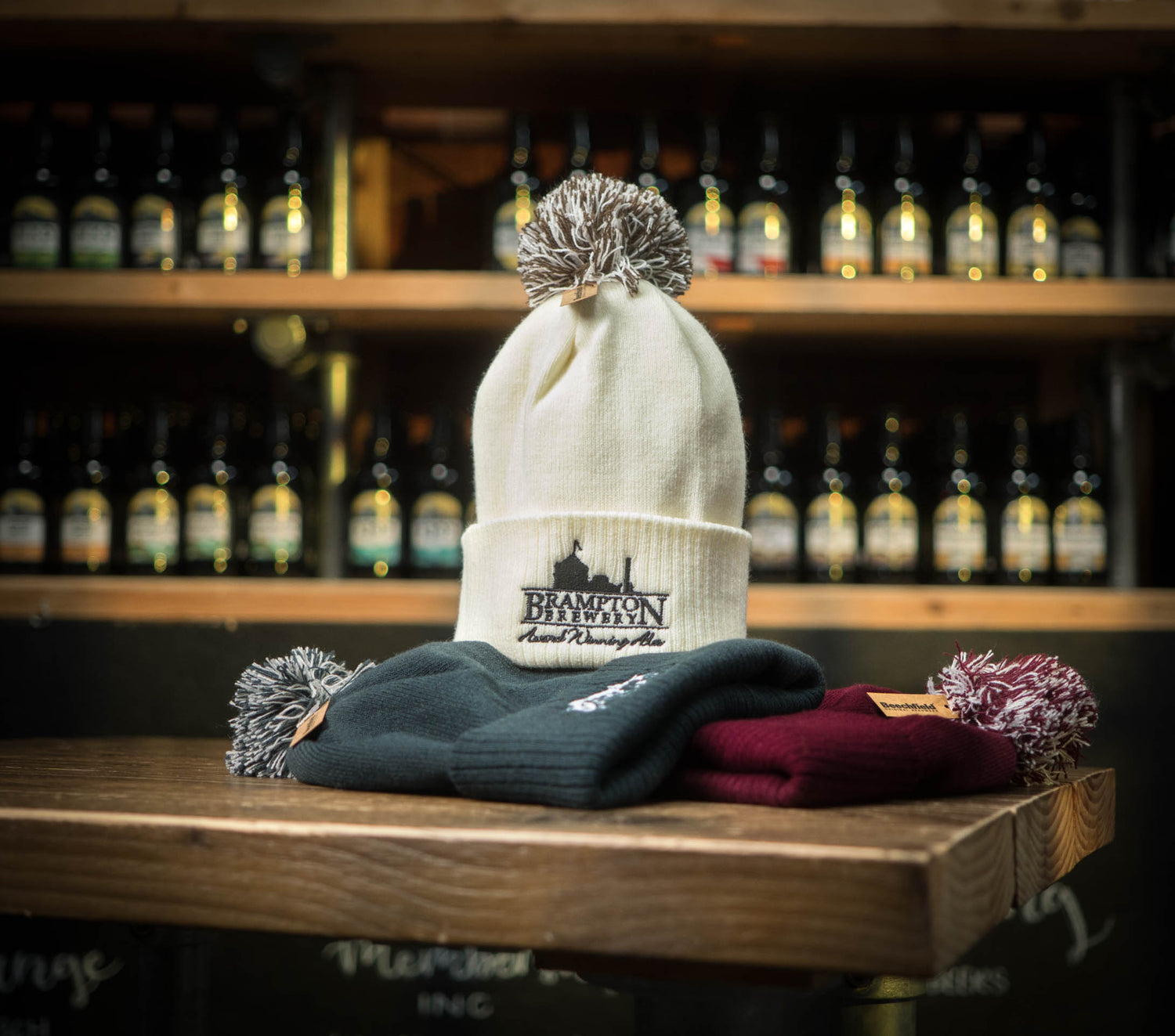 Cream beanie with a brown pom-pom and the Brampton Brewery logo on a wooden surface with a blurred background of bottles.