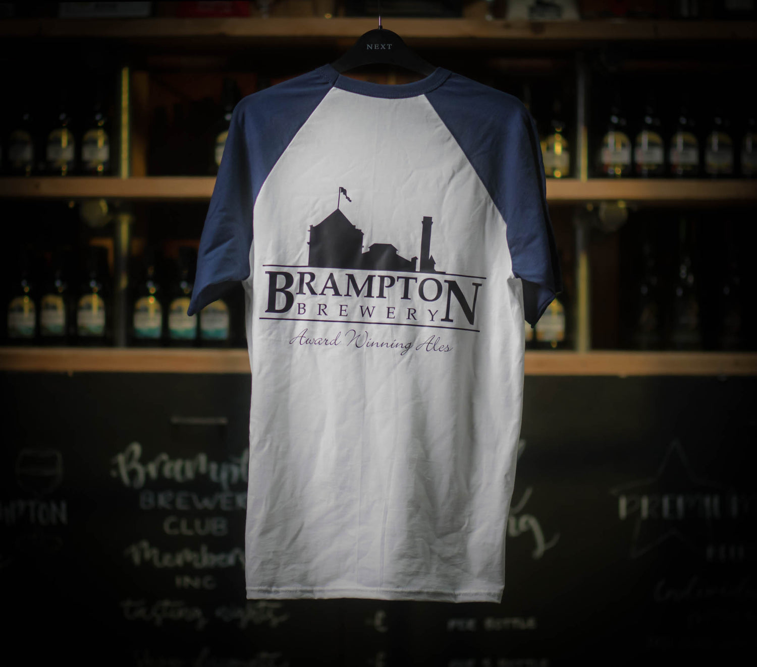 White and navy baseball Brampton Brewery T‑shirt hanging in a rustic brewery setting, featuring the brewery’s logo and “Award Winning Ales” text.