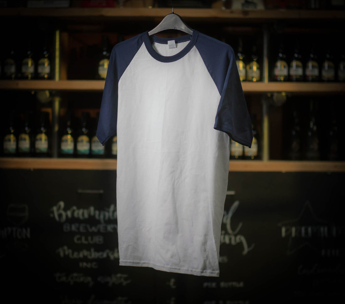 White and navy baseball Brampton Brewery T‑shirt hanging in a rustic brewery setting.