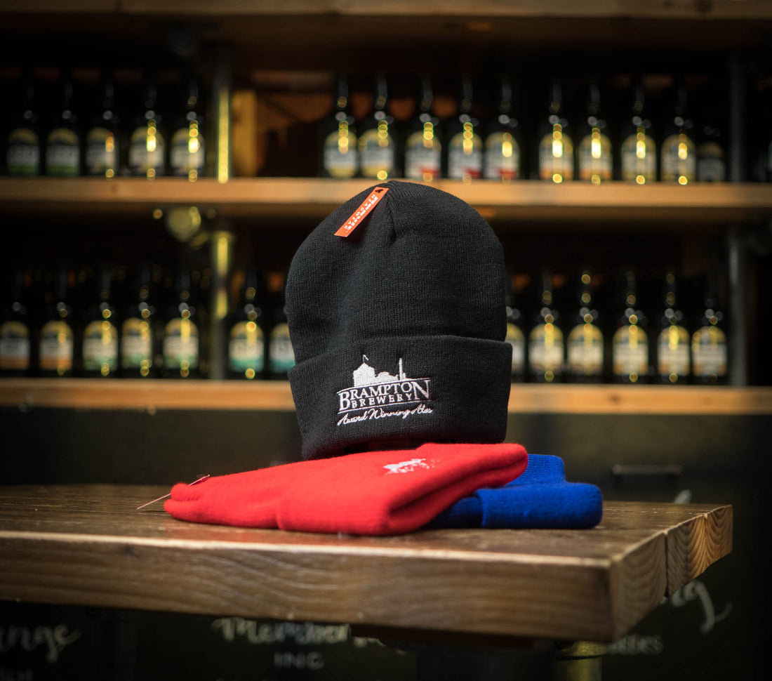 Black beanie with a Brampton Brewery logo on a wooden surface with bottles in the background