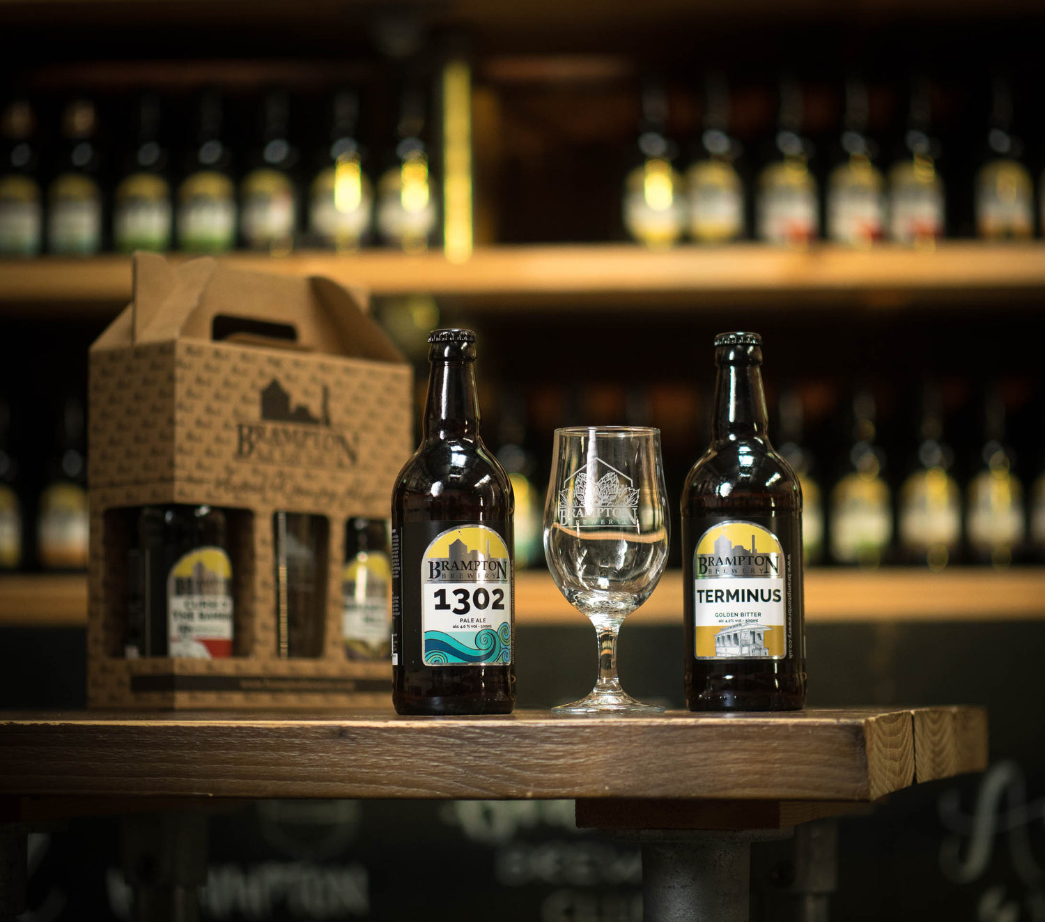 Two bottles of Brampton Brewery beer with a glass on a wooden table, surrounded by more bottles in the background.