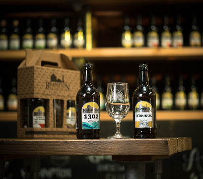 Two bottles of Brampton Brewery beer with a glass on a wooden table, surrounded by more bottles in the background.