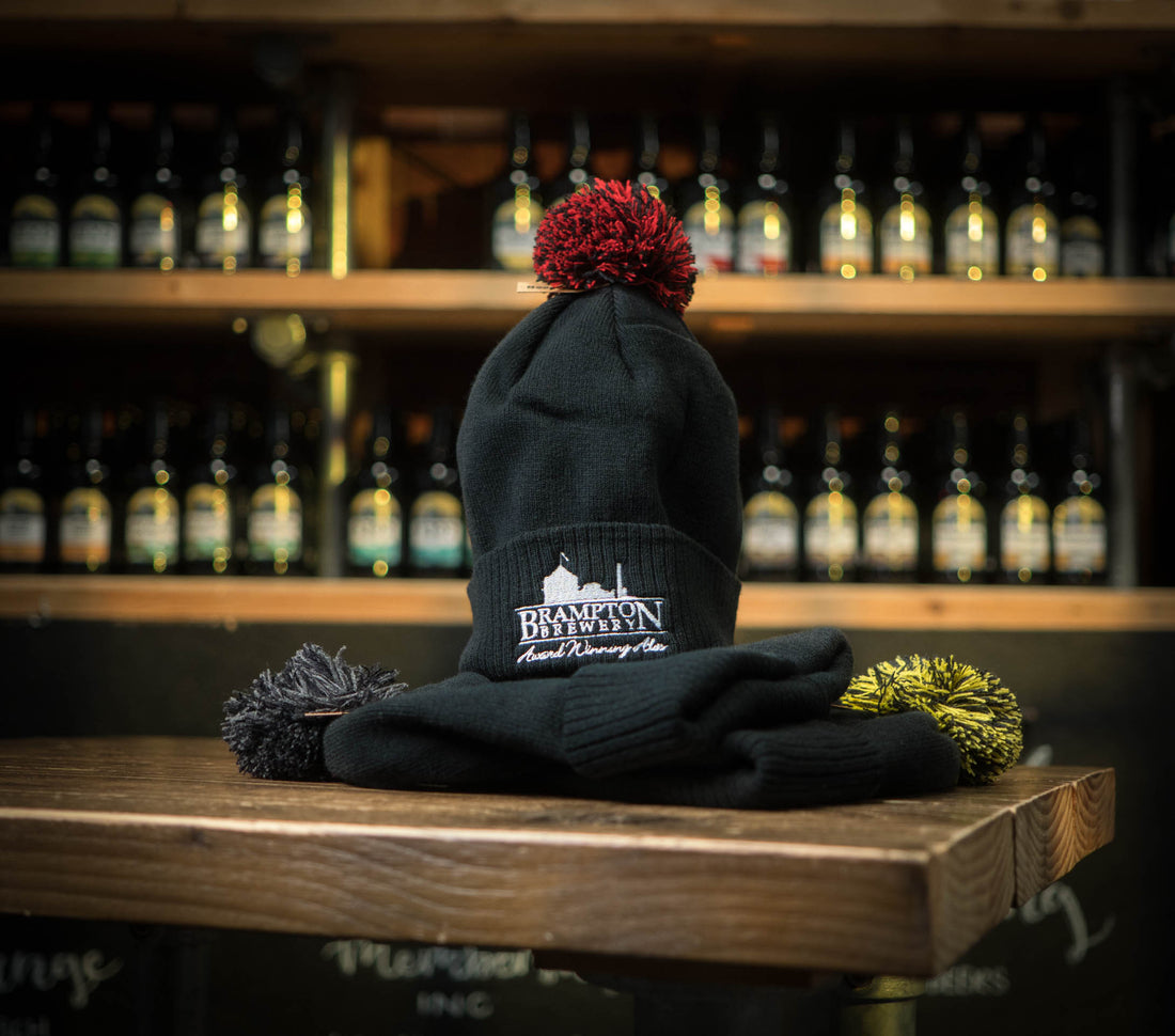 Black beanie with red pom-pom and a Brampton Brewery logo on a wooden surface with bottles in the background