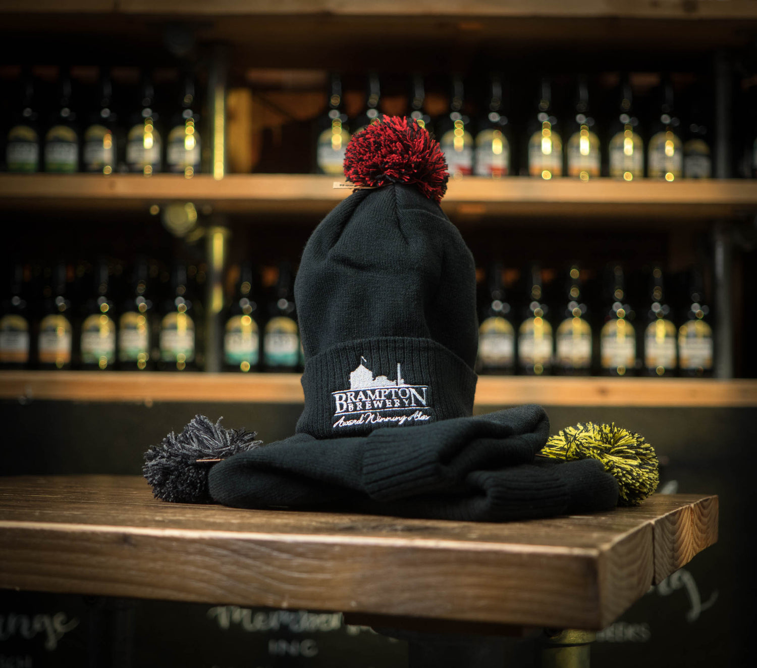 Black beanie with red pom-pom and a Brampton Brewery logo on a wooden surface with bottles in the background