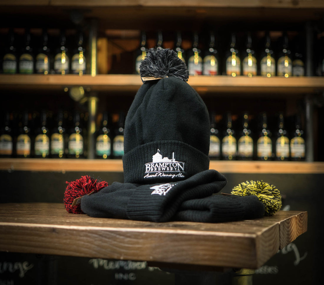 Black beanie with black pom-pom and Brampton Brewery logo on a wooden surface with bottles in the background