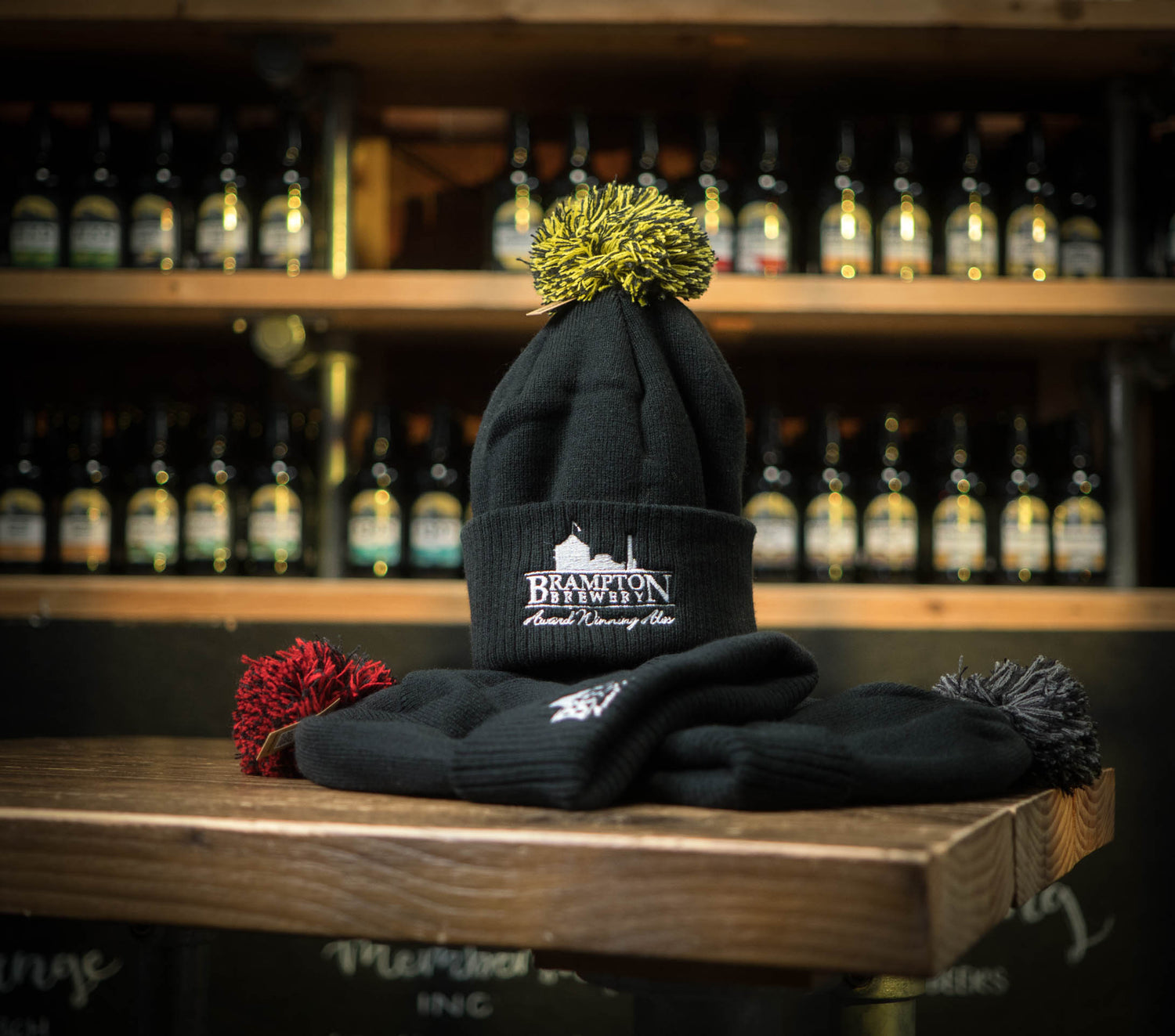 Black beanie with a yellow pom-pom and the Brampton Brewery logo on a wooden surface with a blurred background of bottles.
