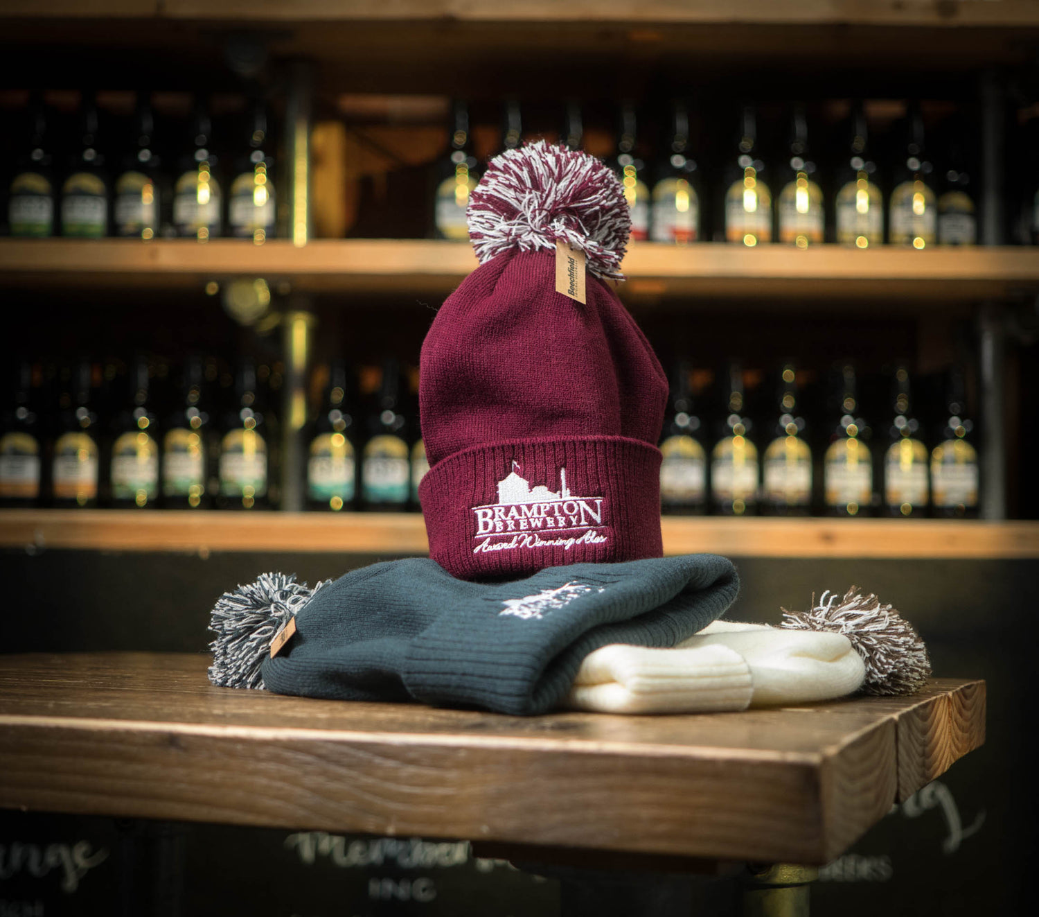Burgundy beanie with a burgundy pom-pom and the Brampton Brewery logo on a wooden surface with a blurred background of bottles.