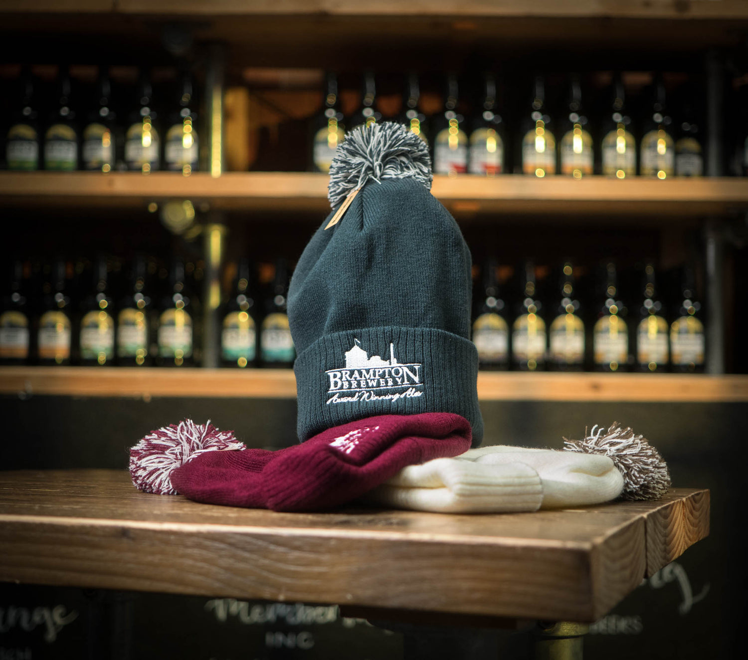Grey beanie with a grey pom-pom and the Brampton Brewery logo on a wooden surface with a blurred background of bottles.
