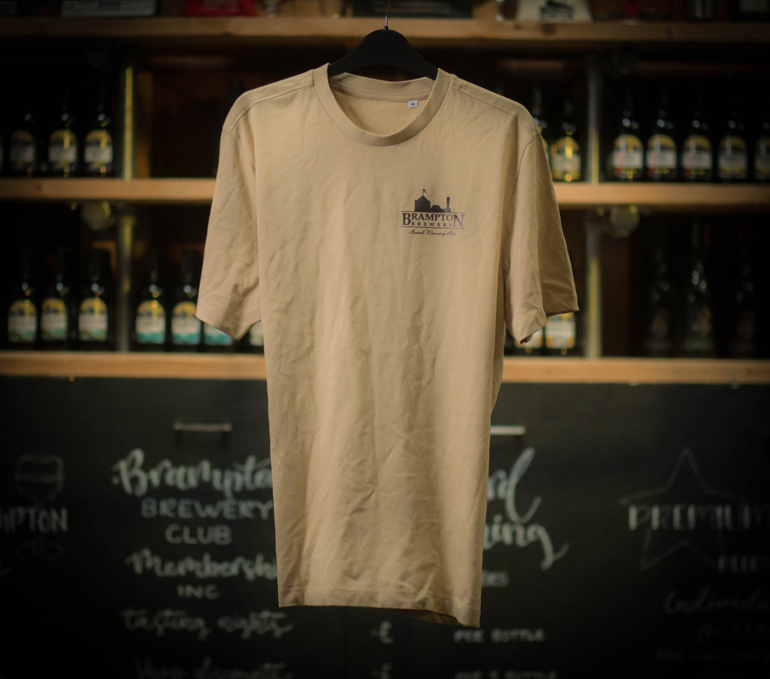 A beige Brampton Brewery T-shirt hanging on a black hanger, featuring a small Brampton Brewery logo printed on the left chest. Shelves of beer bottles and a chalkboard wall appear blurred in the background.