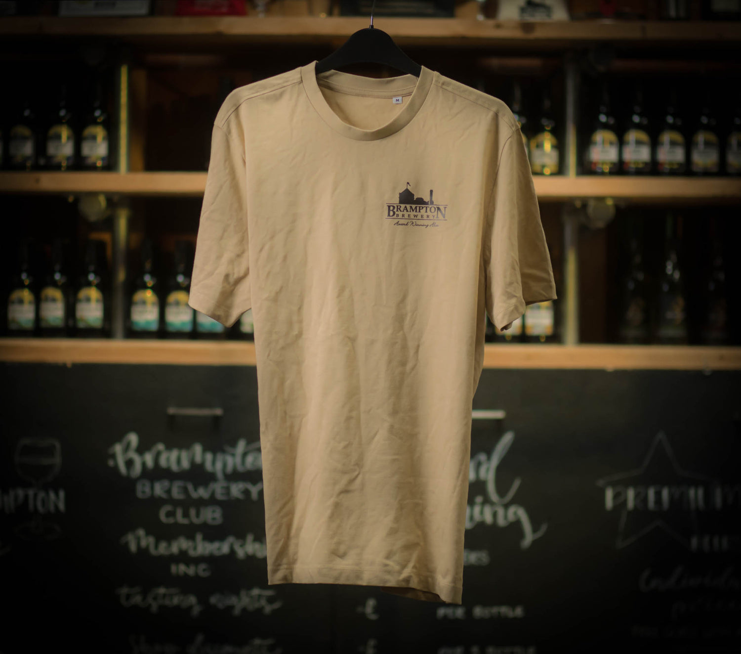 A beige Brampton Brewery T-shirt hanging on a black hanger, featuring a small Brampton Brewery logo printed on the left chest. Shelves of beer bottles and a chalkboard wall appear blurred in the background.