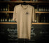 A beige Brampton Brewery T-shirt hanging on a black hanger, featuring a small Brampton Brewery logo printed on the left chest. Shelves of beer bottles and a chalkboard wall appear blurred in the background.