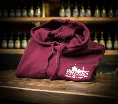 Folded maroon hoodie with a Brampton Brewery logo on a wooden surface with bottles in the background