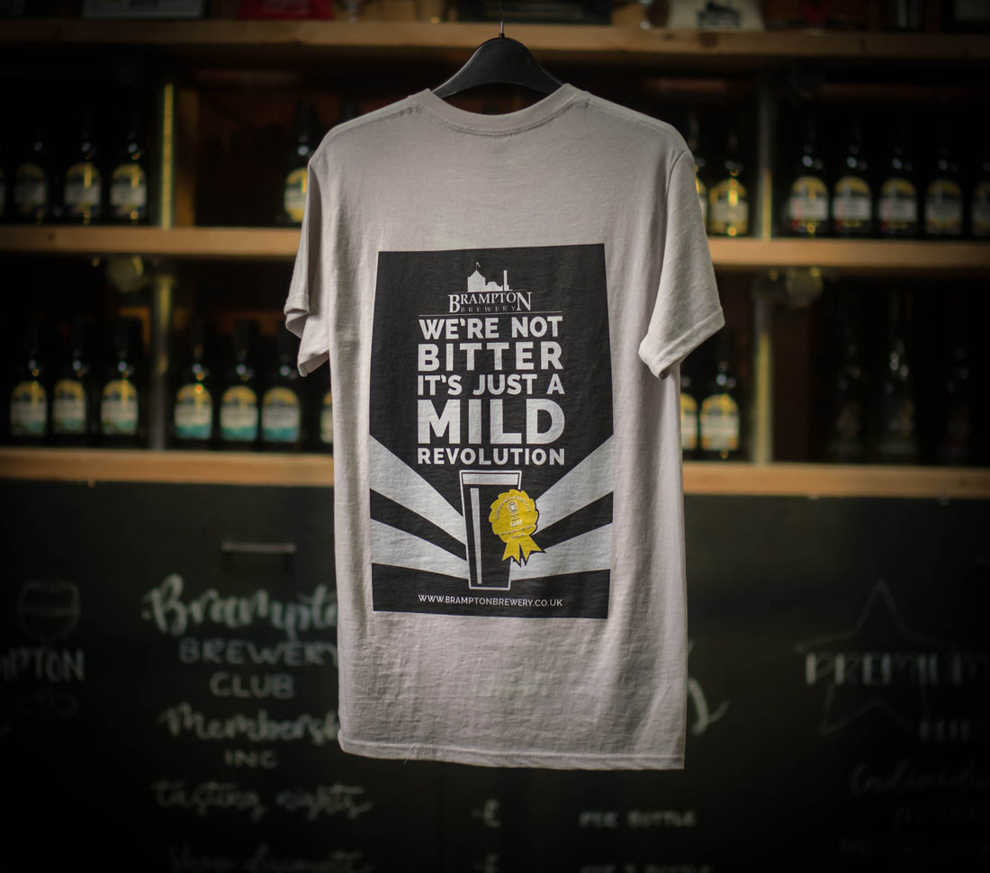 A light grey Brampton Brewery T-shirt hanging on a rail, showing the back print that reads “We’re not bitter, it’s just a mild revolution” above a graphic of a beer glass with a gold award badge. Bottles of beer are displayed blurred in the background.