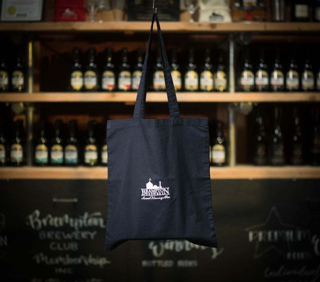 Black tote bag with a Brampton Brewery logo hanging in front of a shelf of bottles.