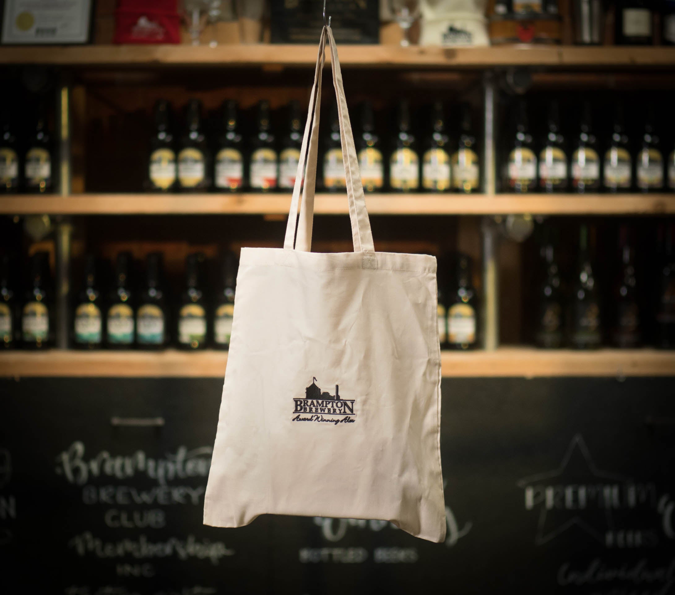 White tote bag with a Brampton Brewery logo hanging in front of a shelf of bottles.