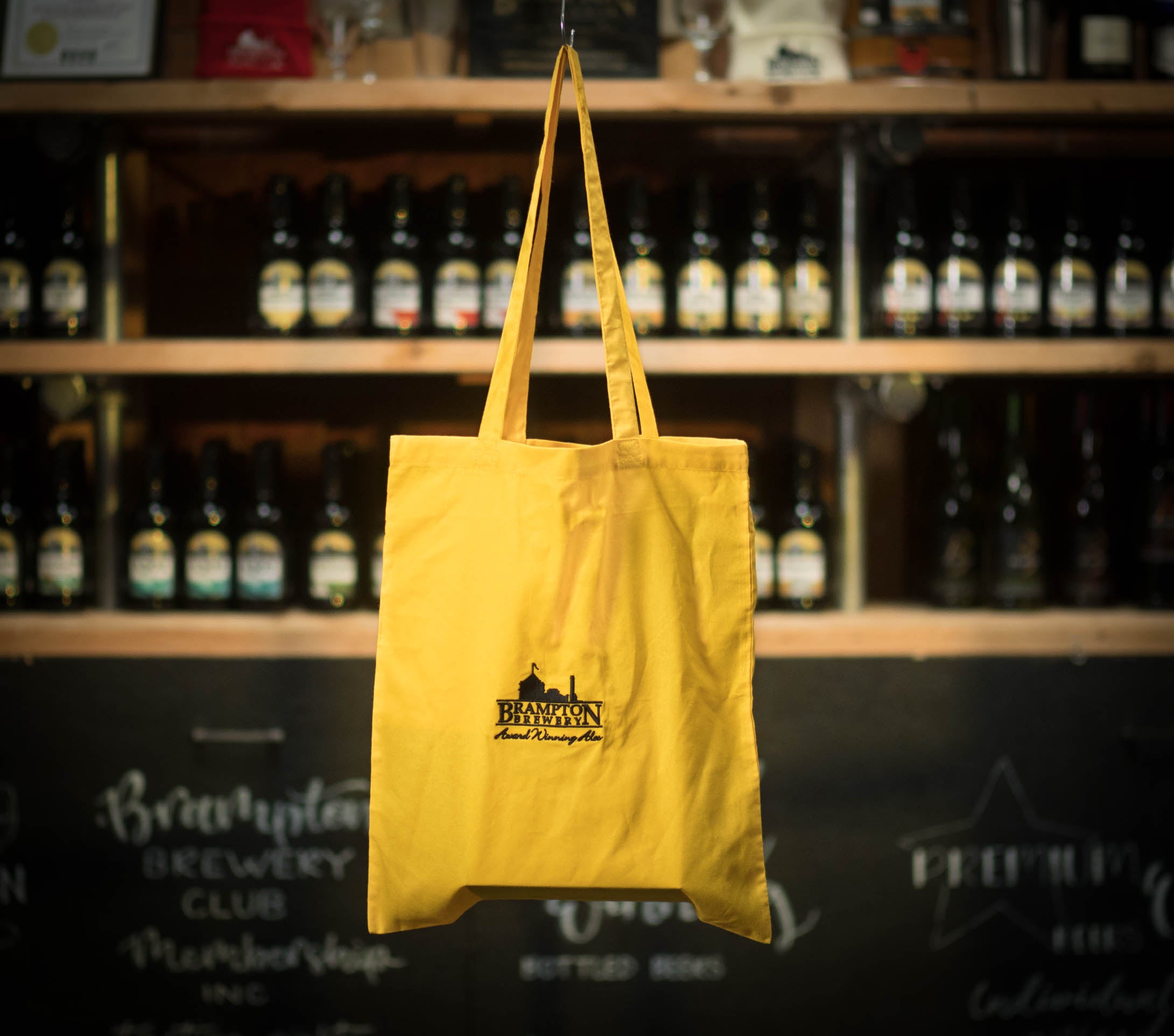 Yellow tote bag with a Brampton Brewery logo hanging in front of a shelf of bottles.